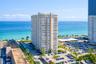 High-rise residential building featuring stacked private balconies, an expansive paved parking area, and mature palm tree landscaping