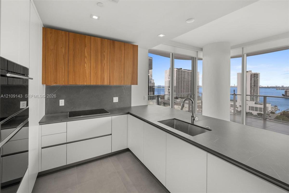Kitchen featuring wood-finish upper cabinetry, matte white base cabinetry, dark stone-finish countertops and backsplash, stainless steel undermount si