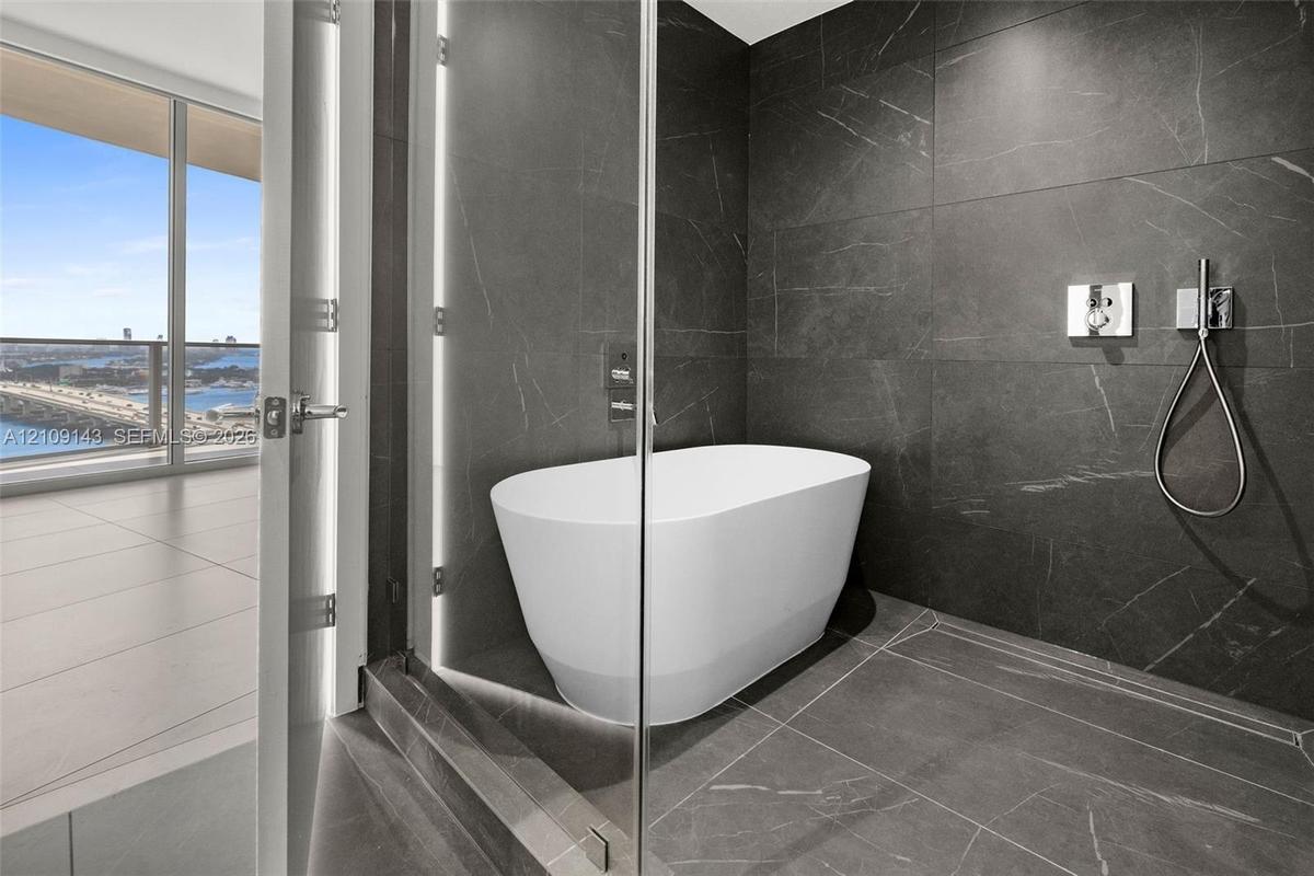 Freestanding soaking tub set within a glass-enclosed wet room featuring floor-to-ceiling dark-toned stone-finish tile and polished chrome plumbing fix