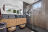 Floating wood-finish vanity with dual sinks, backlit organic-shaped mirror, matte black fixtures, walk-in shower with glass enclosure, and vertical-sl
