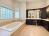 Soaking tub with tile surround, dual-sink vanity with dark cabinetry, light-toned tile flooring, and multi-paned glass block windows