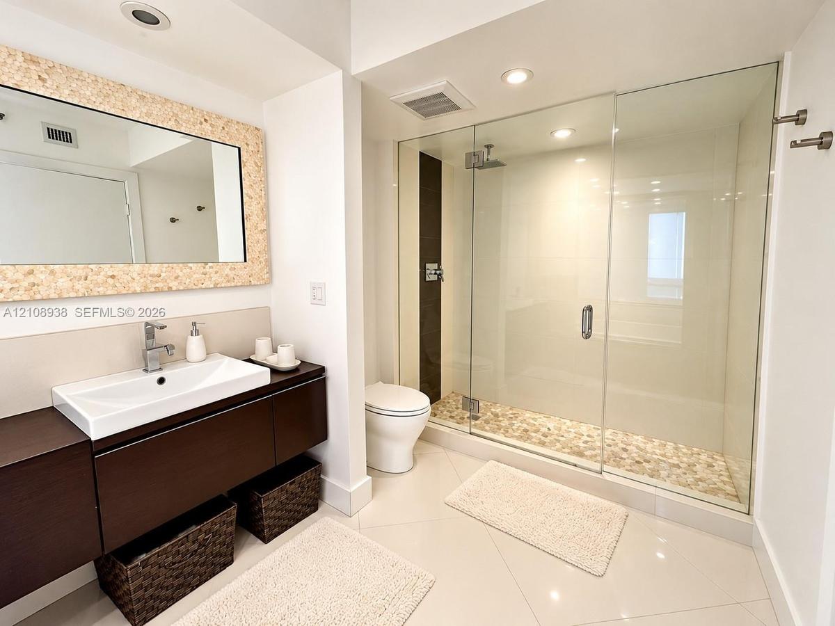 Floating wood-finish vanity with a rectangular vessel sink, frameless glass-enclosed shower featuring a stone pebble floor, and large framed mirror