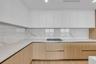 Contemporary kitchen featuring flat-panel upper cabinetry, light-toned wood base cabinetry, stone-surface countertops, full-height stone-surface backs
