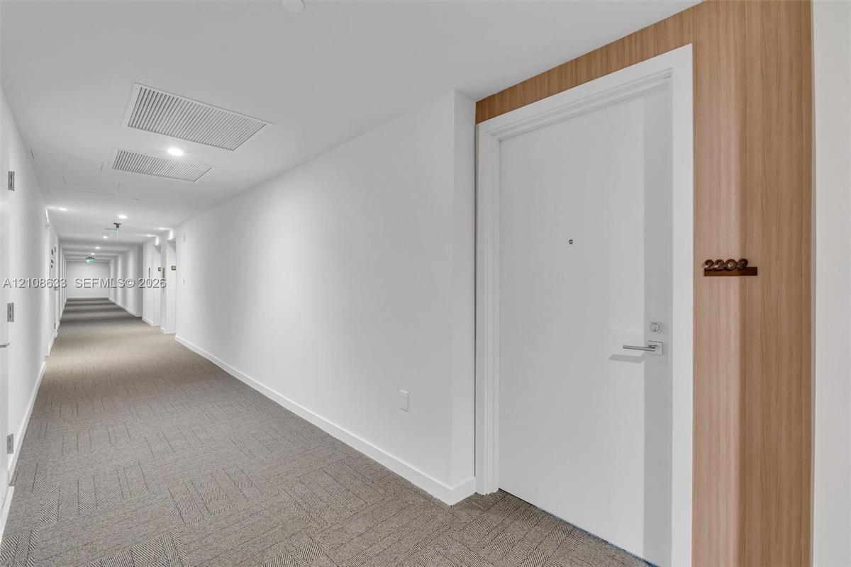 Hallway featuring neutral carpeting, white walls, and a recessed door entry accented with wood-finish paneling and mounted unit signage