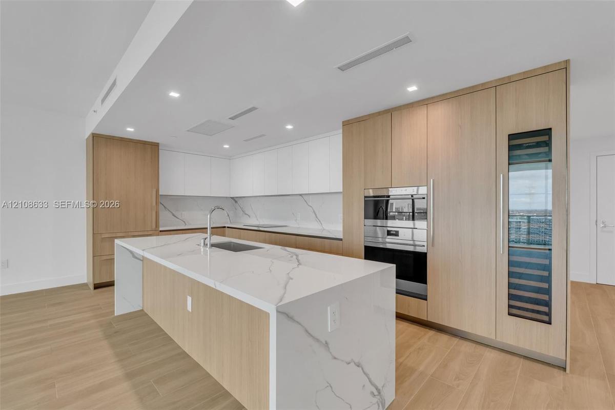 Kitchen featuring wood-finish cabinetry, waterfall-edge island, stone-surfaced countertops and backsplash, integrated oven, and built-in wine refriger