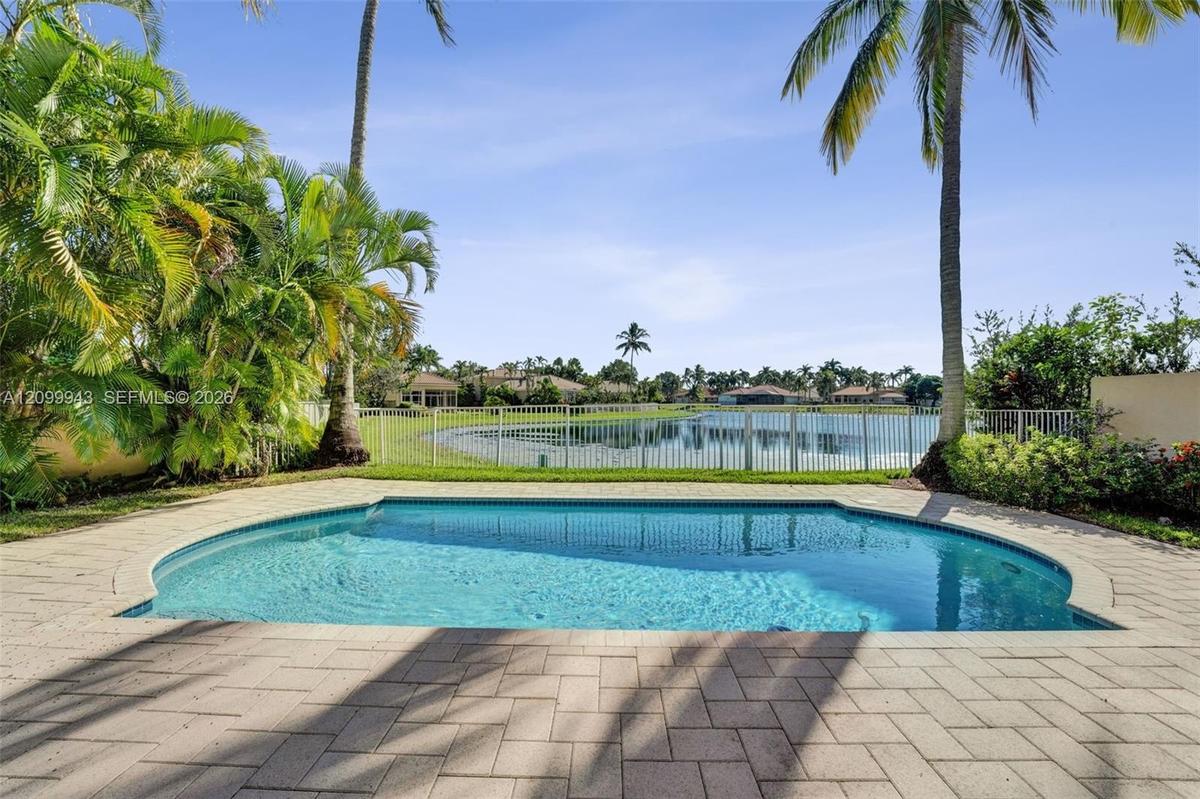 Inground swimming pool with paver decking, enclosed by a perimeter fence, featuring mature palm trees and water frontage