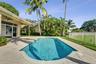 In-ground swimming pool with a stone paver deck, covered patio, white metal fencing, and mature palm trees