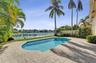 In-ground swimming pool surrounded by a paved stone patio, metal perimeter fencing, waterfront location, and mature palm trees