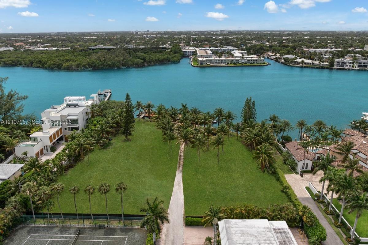 Waterfront lot featuring direct canal access, expansive manicured lawn, mature palm trees, and a hard-surface tennis court