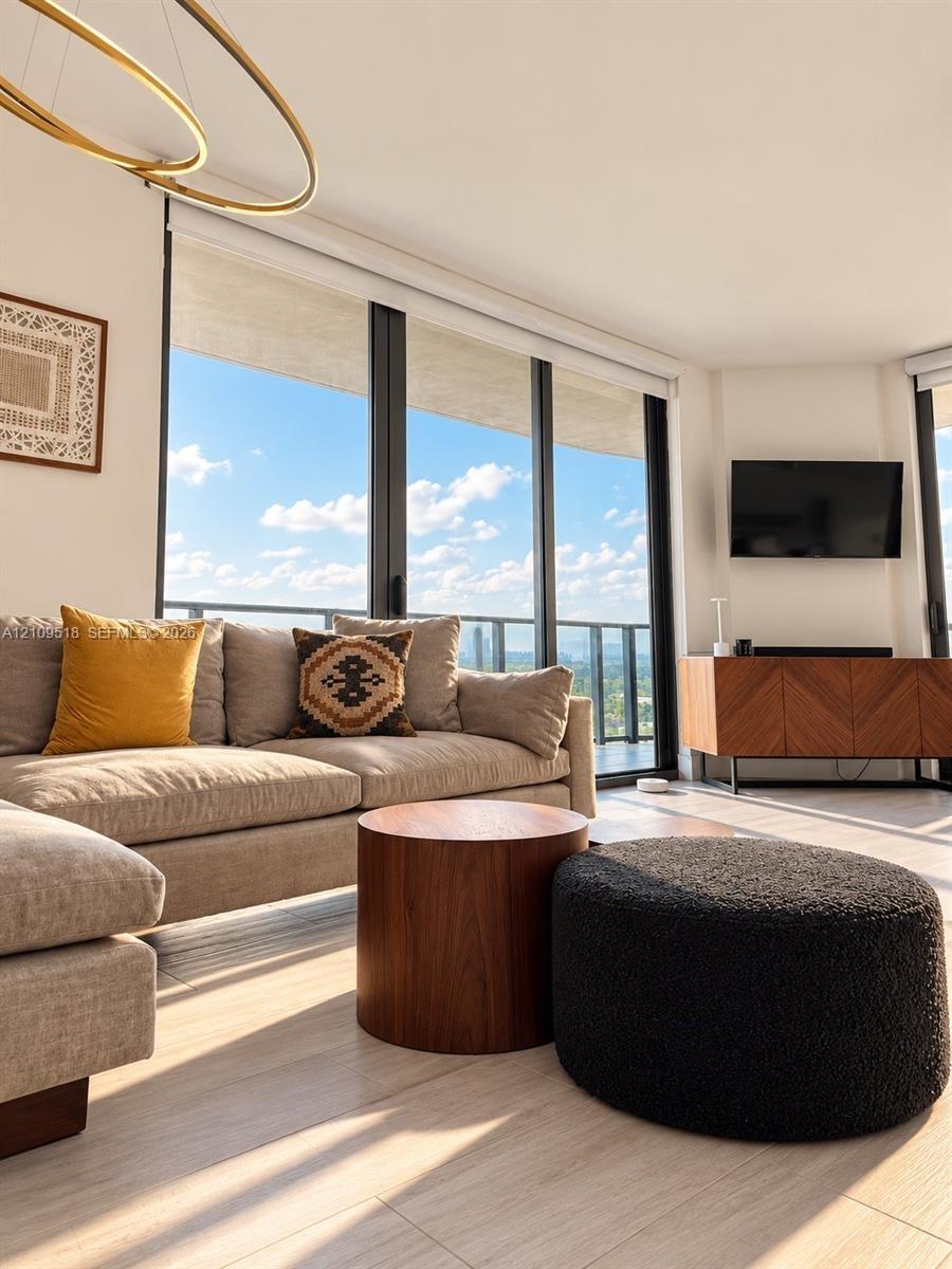 Floor-to-ceiling glass paneling with integrated balcony access, neutral stone-finish flooring, gold-tone modern circular chandelier, and custom built-