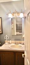 Vanity featuring wood-finish cabinetry, a white integrated countertop and sink, a three-light decorative wall fixture, and a rectangular mirror