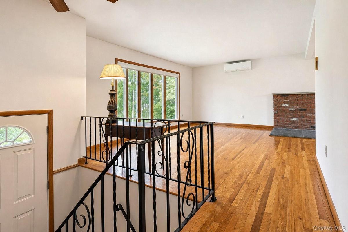 Hard wood flooring, brick fireplace surround, decorative wrought iron railing, expansive picture window, and wall-mounted climate control unit part of