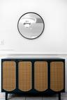 Circular wall mirror with a matte black frame, set above a built-in dark cabinetry unit featuring woven rattan door panels and brass-finish hardware