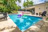 Swimming pool with stone paver decking and covered patio area featuring a ceiling fan, surrounded by privacy fencing and mature palm trees