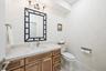 Natural-finish wood cabinetry, stone-surface countertop with an integrated sink, polished metal faucet, decorative mirror frame, and textured wallpape
