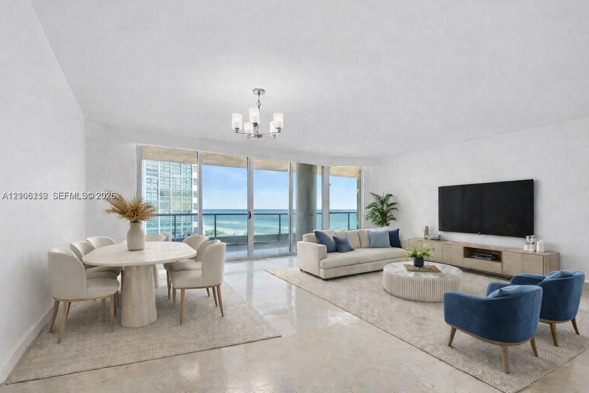 Open-concept interior featuring light-toned stone flooring, floor-to-ceiling glass sliding doors, a glass-railed balcony, and a multi-arm chandelier