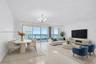 Open-concept interior featuring light-toned stone flooring, floor-to-ceiling glass sliding doors, a glass-railed balcony, and a multi-arm chandelier