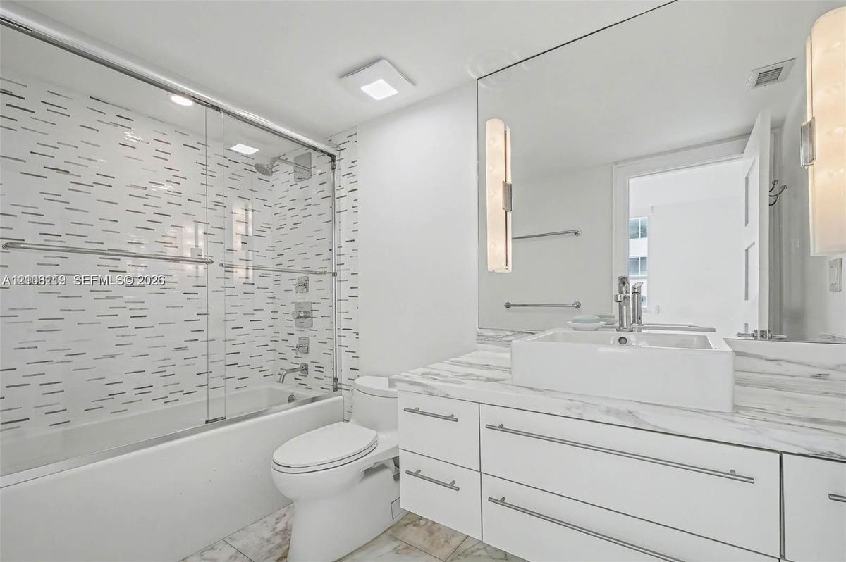 Bathroom featuring a white vanity with a stone-finish countertop and square vessel sink, full-width wall mirror with integrated lighting, glass-enclos
