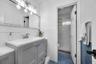 Grey vanity with white countertop, integrated sink, polished metal faucet, multi-bulb vanity light, and white tiled wall surround