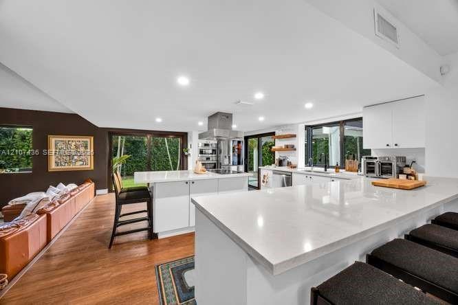 Modern kitchen featuring an expansive central island with quartz countertops, custom white cabinetry, stainless steel range hood, recessed lighting, a