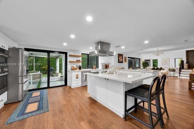 Kitchen featuring a large central island with light-toned solid surface countertops, white cabinetry, stainless steel range hood, built-in wall oven, 