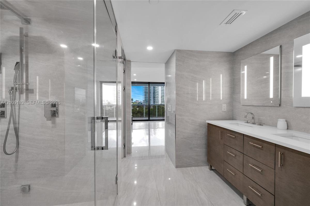 Primary Bathroom featuring large-format tile wall cladding, a double vanity with stone countertop, frameless glass shower enclosure with rainfall show