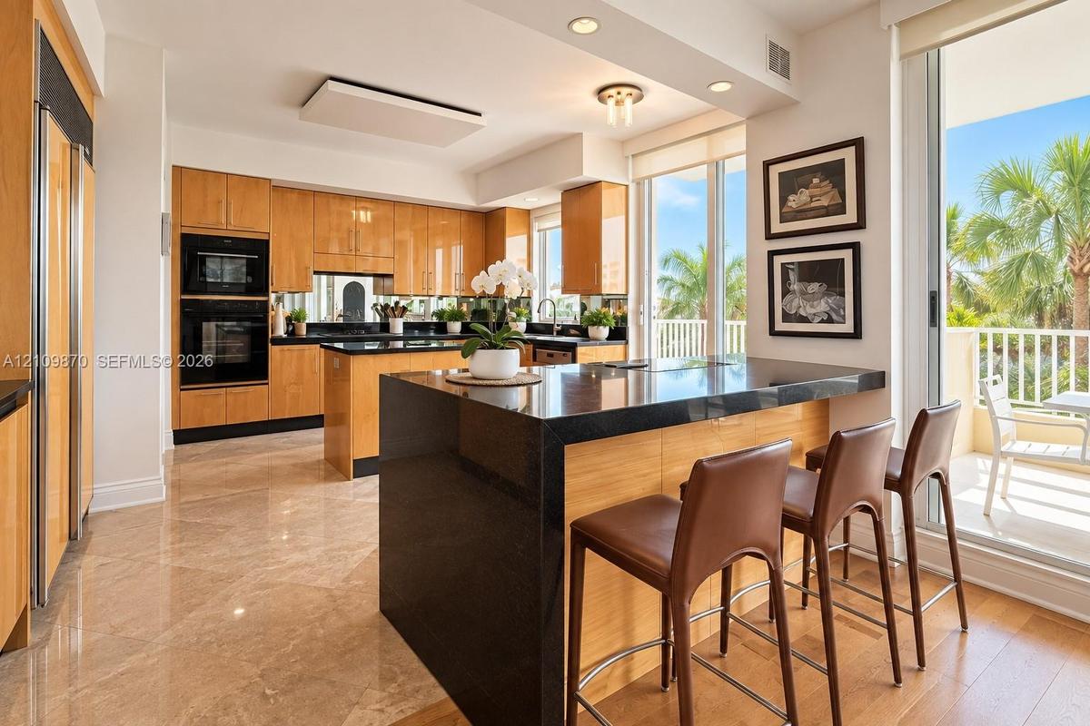 Kitchen featuring wood cabinetry, integrated appliances, dark stone countertops, an expansive island with bar seating, and stone flooring