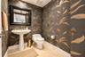 Powder room featuring patterned wallpaper, white pedestal sink, brass-finish widespread faucet, framed wall mirror, and natural stone tile flooring