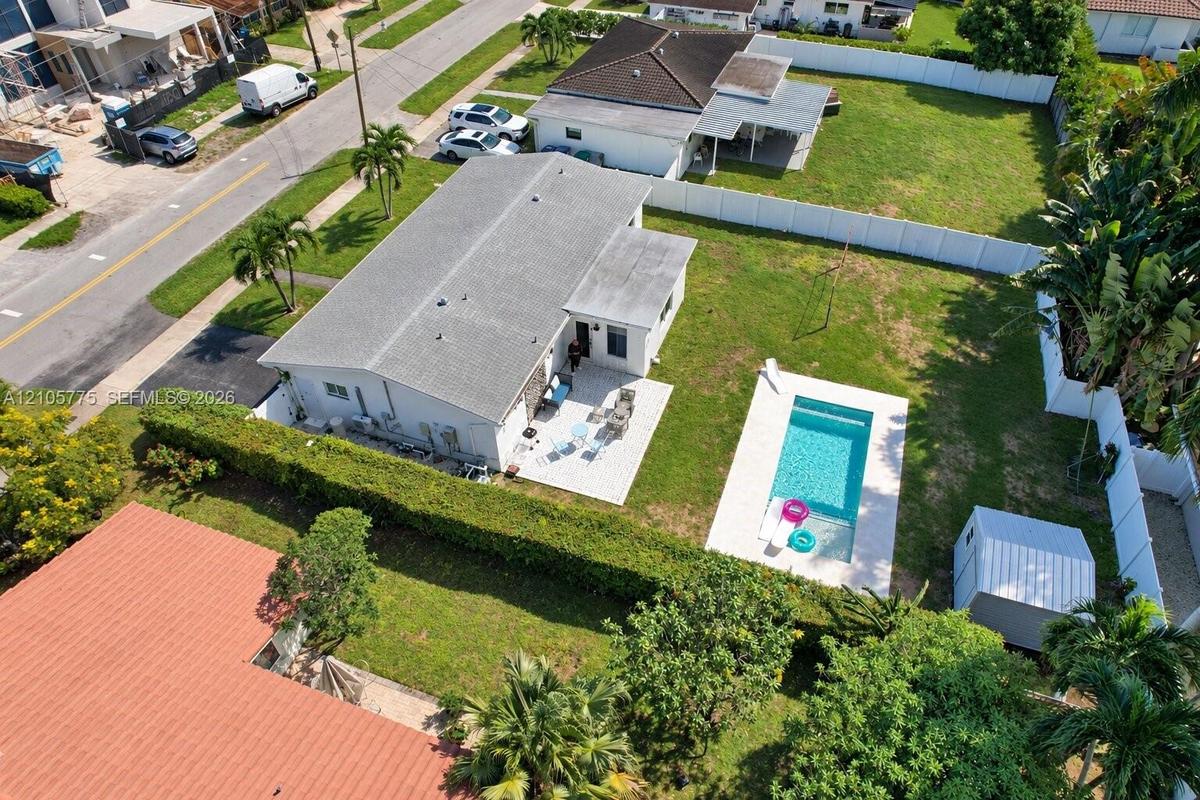 Aerial perspective of a property featuring an in-ground swimming pool, paved patio, expansive lawn, white vinyl perimeter fencing, and a detached stor