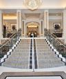 Grand dual staircase featuring intricate wrought iron railings, stone flooring with decorative inlay, and a central crystal chandelier under vaulted c