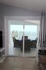 Sliding glass door opening to a covered balcony with a white metal railing, tile flooring, and coastal water backdrop
