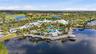 Aerial view of a coastal-inspired complex featuring a resort-style swimming pool, expansive waterfront sun deck, multi-tiered white roofing, and manic