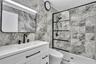 Bathroom featuring full-height stone-finish tile surround, white vanity with quartz countertop, matte black fixtures, framed vanity mirror, and glass 
