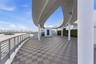 Expansive outdoor terrace featuring a geometric tile deck, curved architectural overhang, cylindrical support columns, metal railing, and privacy hedg