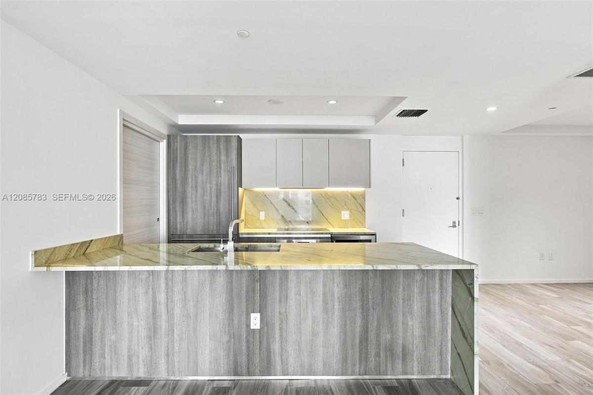 Kitchen featuring flat-panel wood-finish cabinetry, stone slab countertops, a waterfall-edge island with an integrated sink, under-cabinet lighting, a