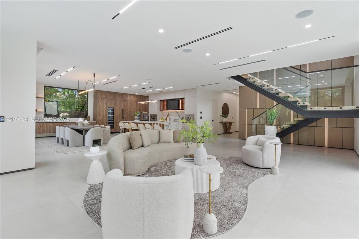 Open-concept floor plan featuring expansive stone-tile flooring, recessed linear lighting, custom wood-paneled cabinetry, and a floating staircase wit
