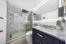 Modern vanity with navy cabinetry, stone countertop, and brass hardware, featuring a glass-enclosed shower with floor-to-ceiling stone tiling, overhea