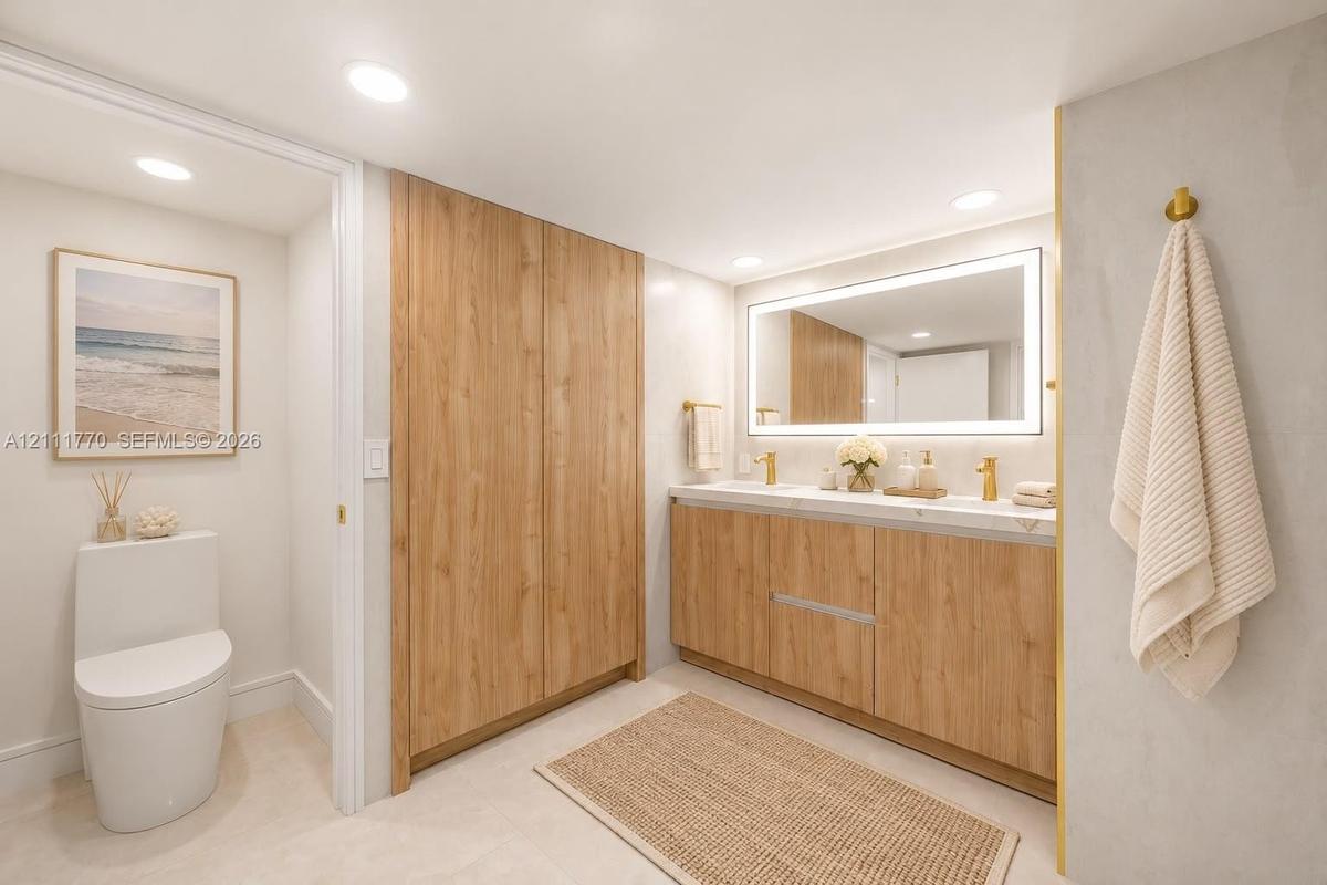 Bathroom featuring a wood-finish double vanity with dual gold-toned faucets, integrated solid-surface countertop, full-width illuminated vanity mirror