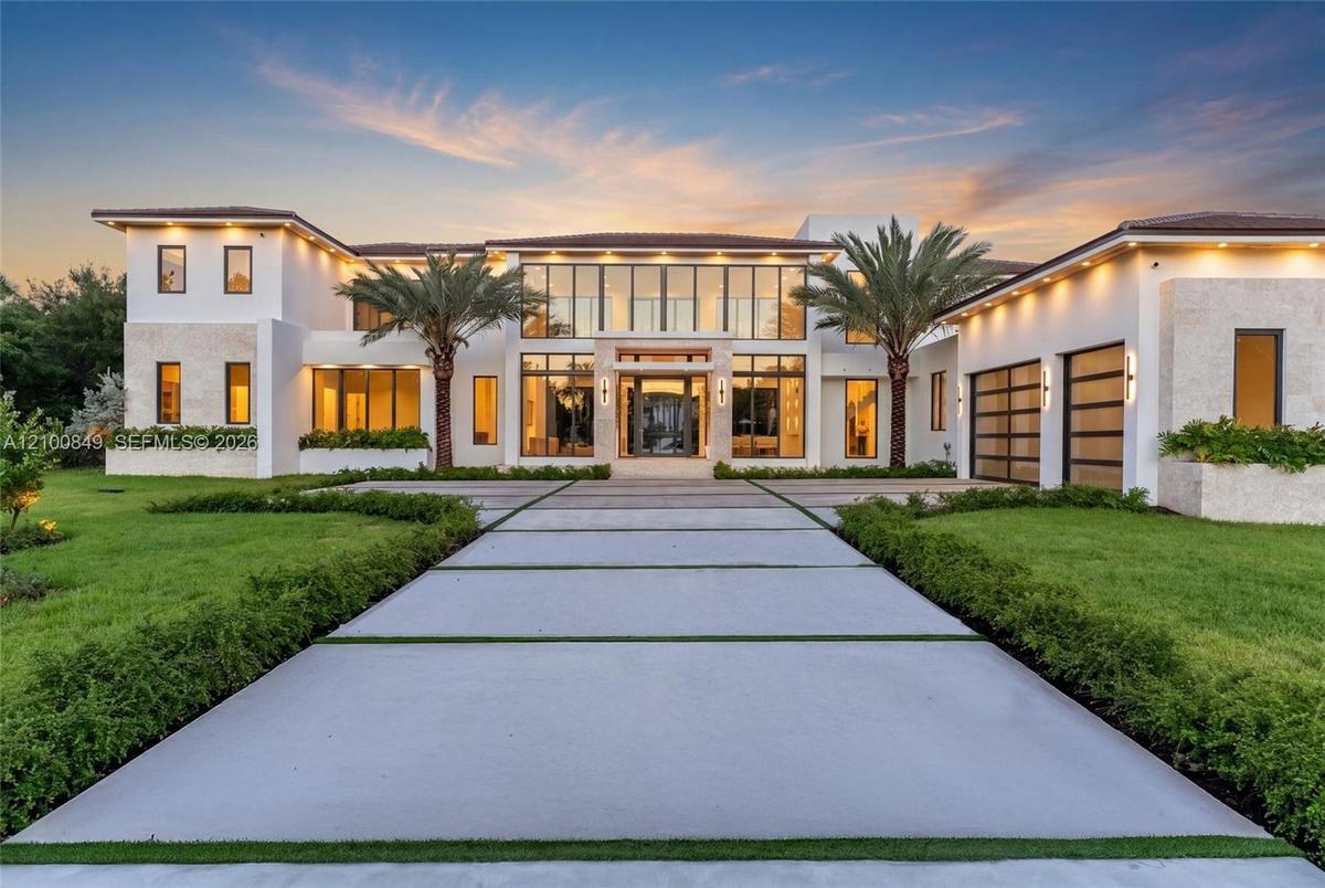 Modern two-story estate featuring a smooth stucco finish, extensive floor-to-ceiling glass wall facade, paved driveway with geometric grass inlay, til
