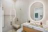 Bathroom featuring a floating wood-finish vanity with a stone countertop, integrated sink, illuminated arched mirror, and a walk-in shower with floor-