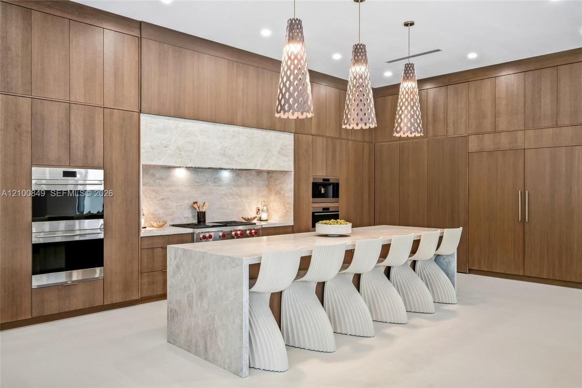 Custom floor-to-ceiling wood cabinetry, expansive stone-topped kitchen island, dual built-in ovens, stone slab backsplash, and three architectural pen