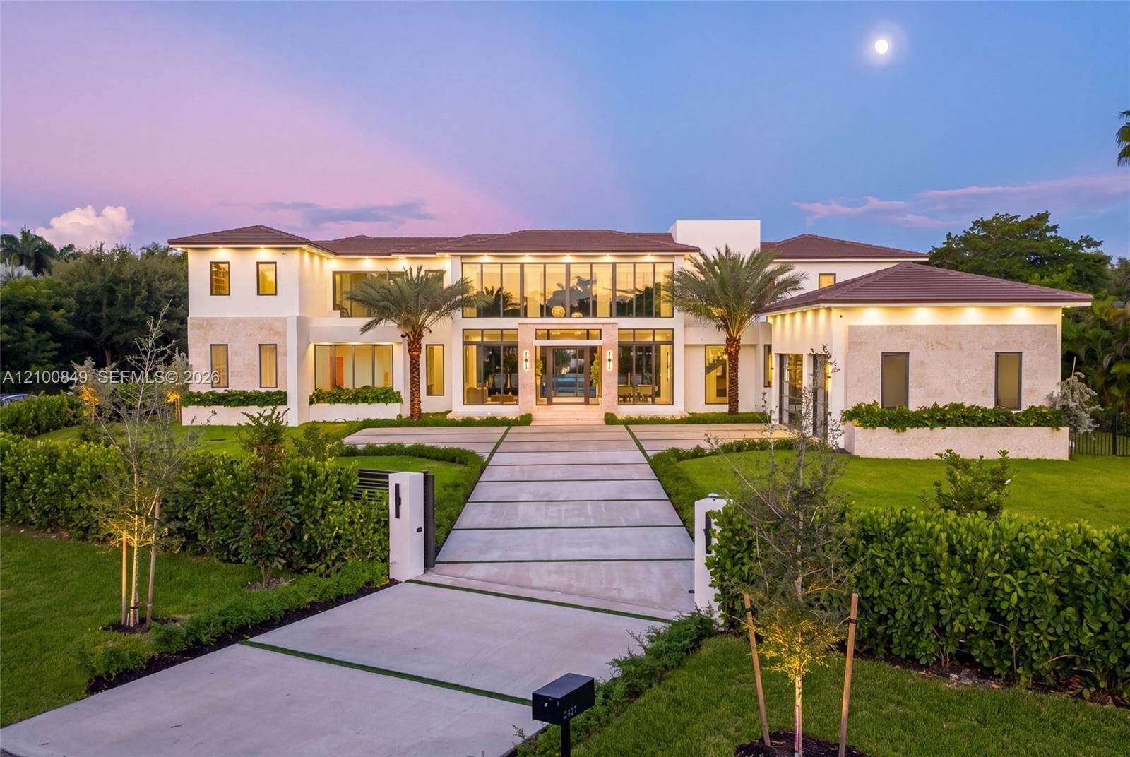 Modern residence featuring a light-toned stone facade, floor-to-ceiling multi-panel windows, a tile roof, and an expansive concrete paver driveway