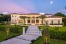 Modern residence featuring a light-toned stone facade, floor-to-ceiling multi-panel windows, a tile roof, and an expansive concrete paver driveway
