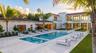 Custom pool with built-in stepping stones, expansive stone-paved patio, two-story white stucco exterior, multiple glass balcony railings, and lush per