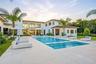 Two-story stucco exterior with a multi-level roofline, featuring a swimming pool with a built-in sun shelf, stone-paver pool deck, and integrated land