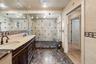 Dual-sink vanity with ornate wood cabinetry, stone countertops, walk-in glass shower enclosure with built-in bench, and patterned stone tile flooring