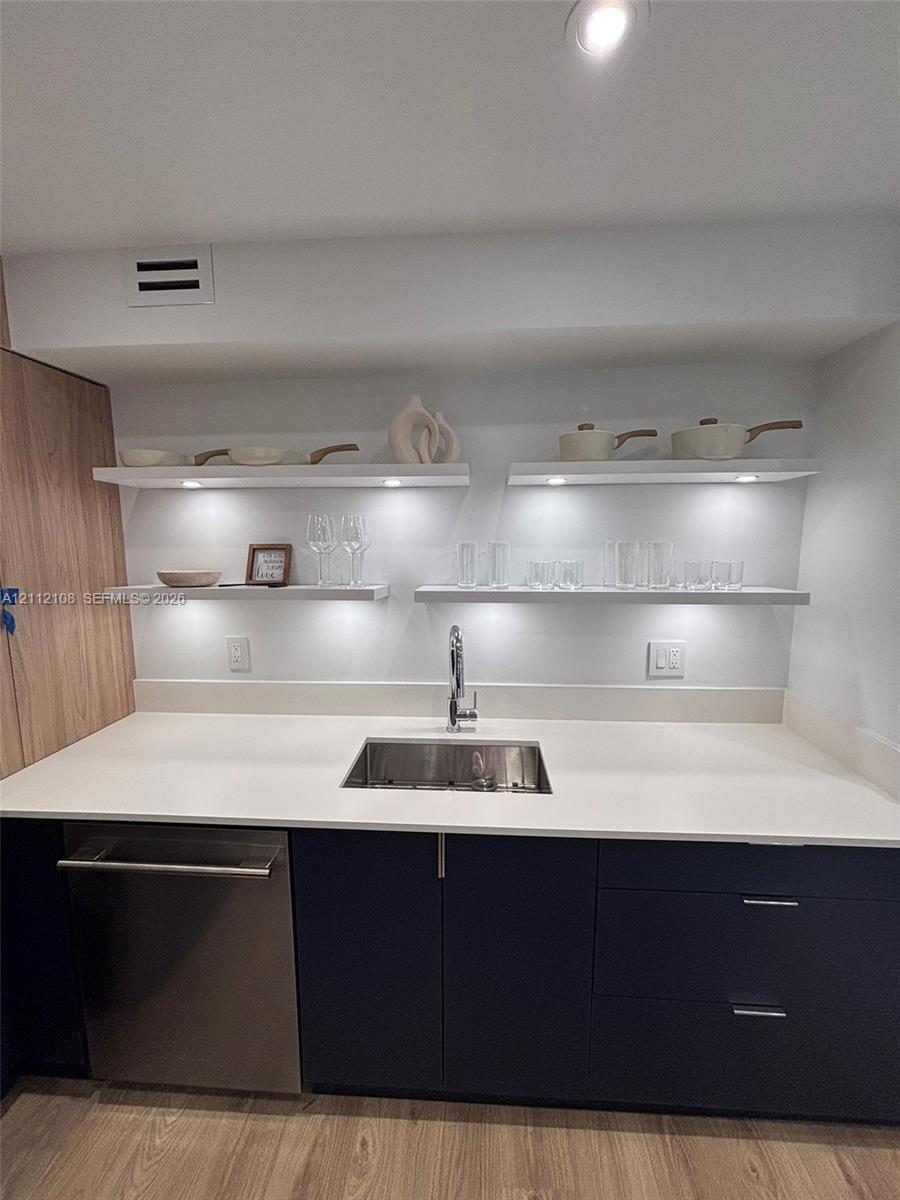 Kitchenette area featuring flat-panel dark cabinetry, integrated stainless steel dishwasher, solid-surface countertop, recessed under-shelf lighting, 