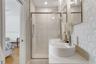 Dual vessel sinks with polished chrome faucets, quartz-surfaced vanity, glass-enclosed shower with recessed shelving, and decorative geometric wall ti
