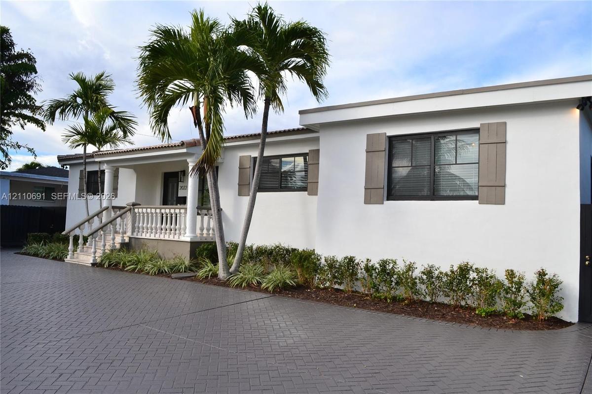 Stucco exterior featuring a covered front porch with a concrete balustrade, herringbone-patterned paved driveway, decorative shutters, and mature palm
