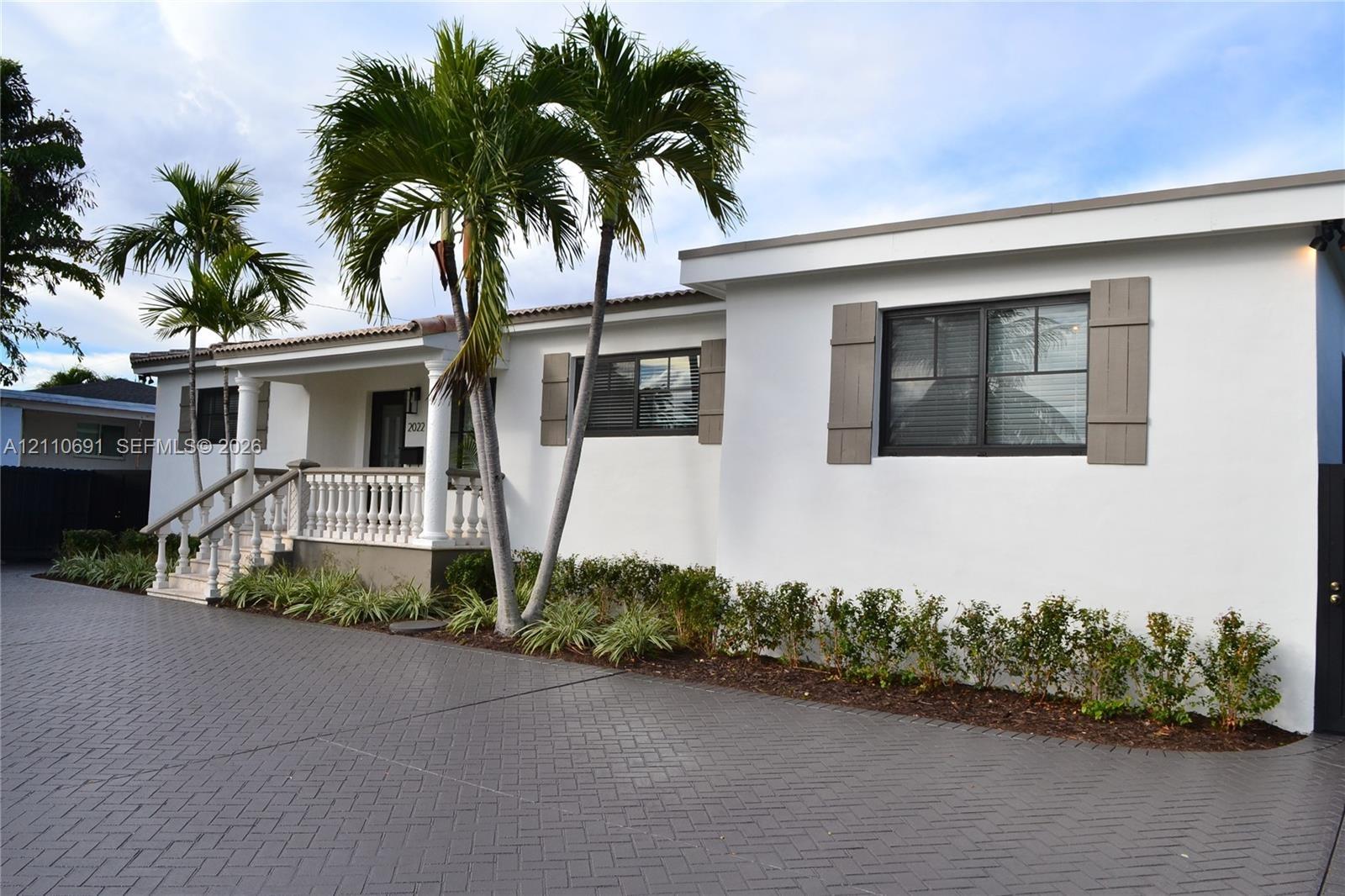 Stucco exterior featuring a covered front porch with a concrete balustrade, herringbone-patterned paved driveway, decorative shutters, and mature palm